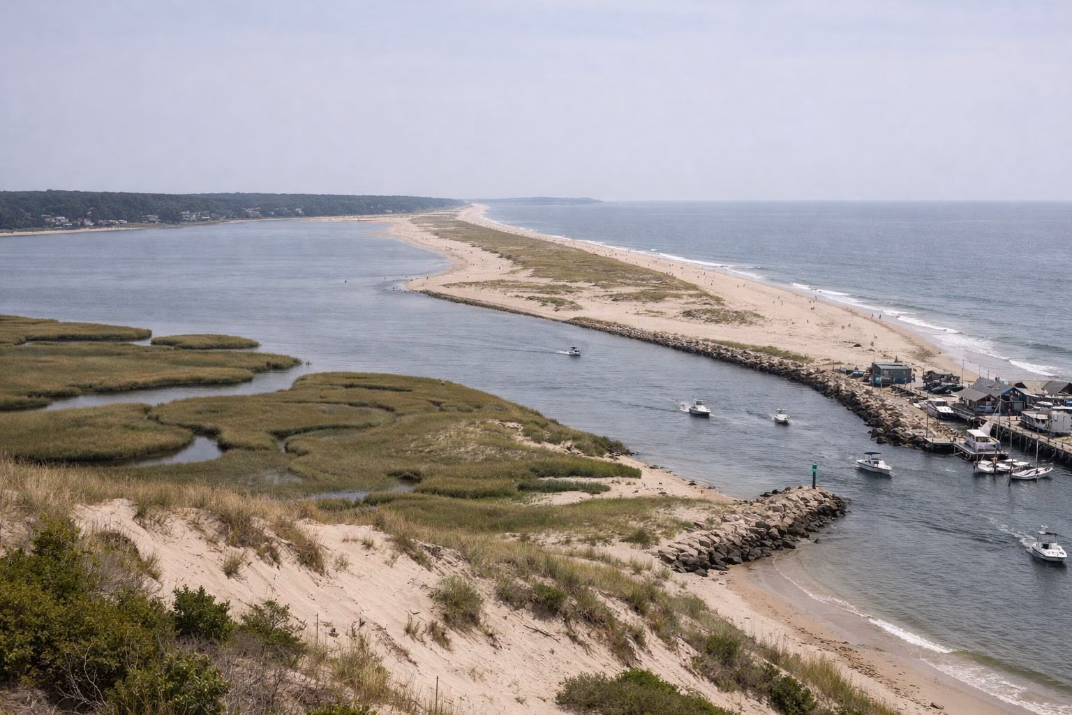 An elevated view of a barrier beach, salt marsh and a river mouth with boats in the channel and a stone jetty