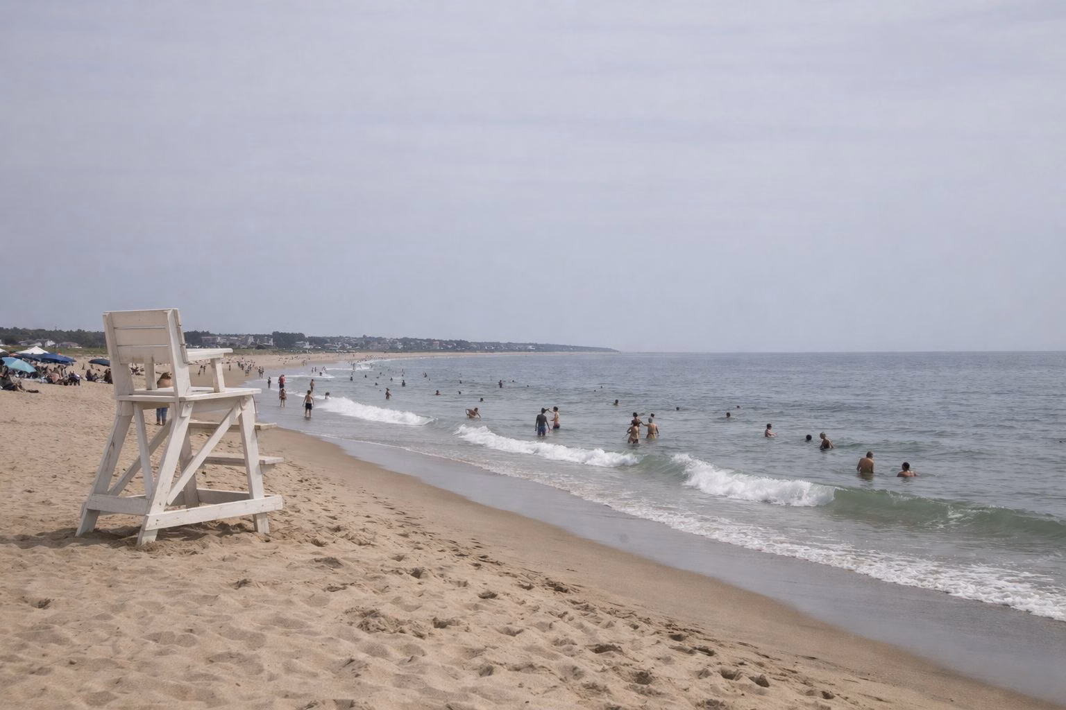 Swimmers in gentle surf along a long sandy beach on a hazy summer day, a white lifeguard chair on the sand