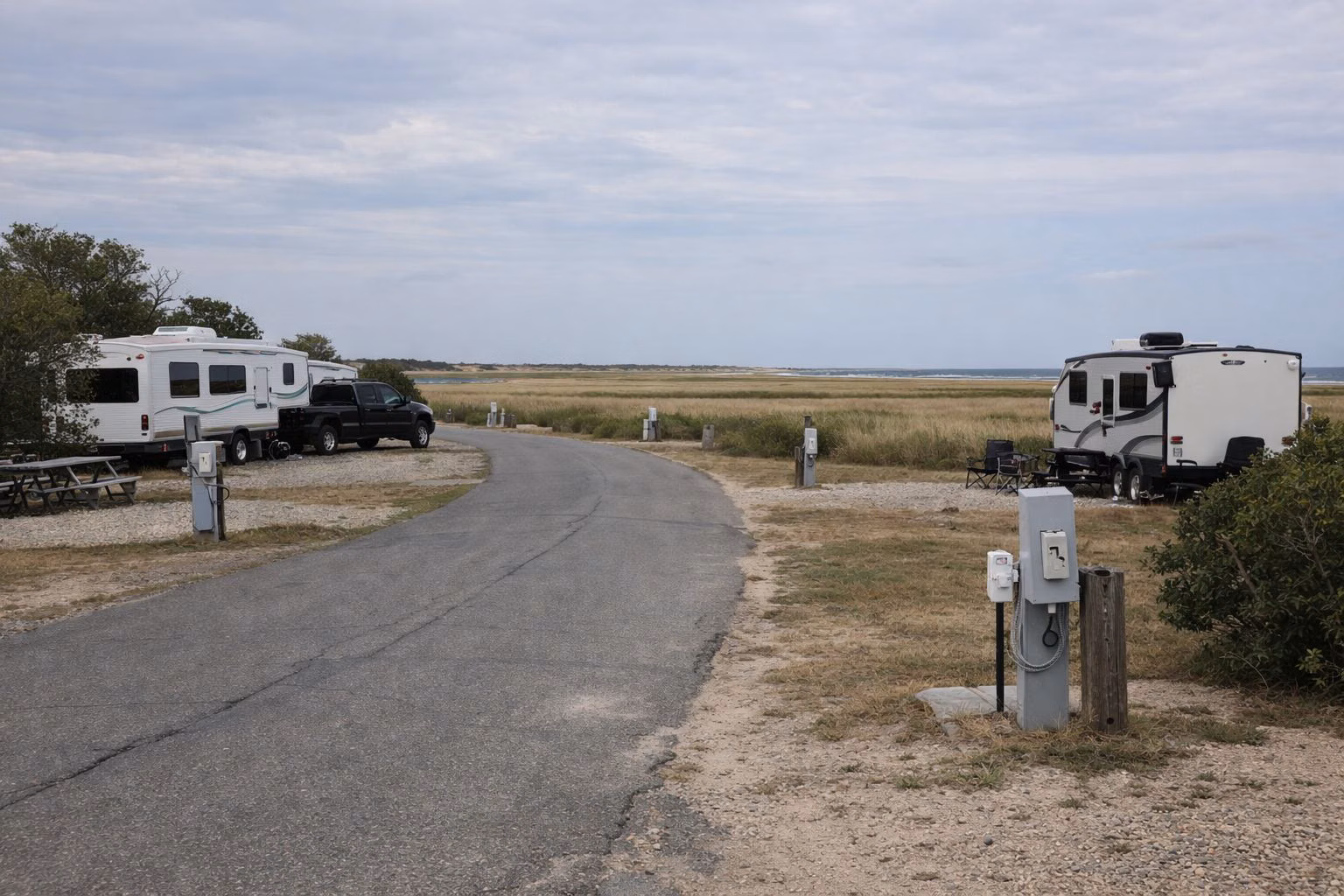 A paved campground loop road past trailer sites with electric hookup posts, salt marsh and the sea beyond