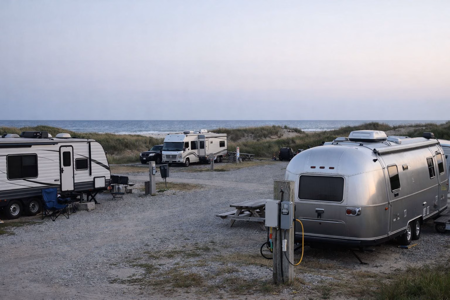 Travel trailers and RVs on gravel campsites with an electric hookup post, low dunes and the ocean behind