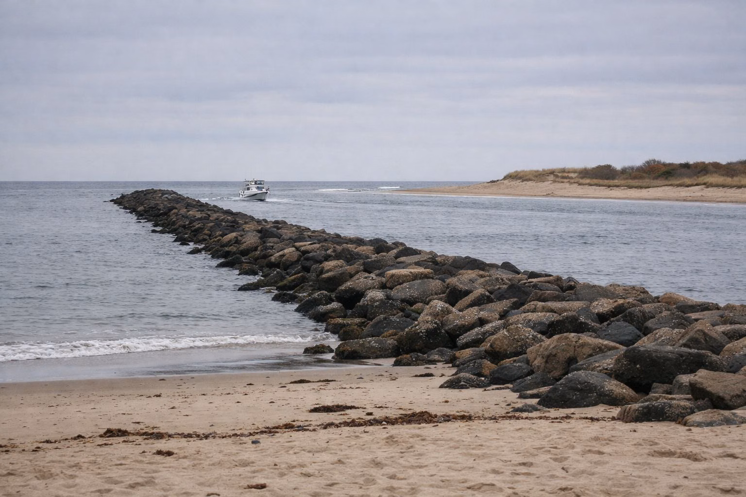 A long low rock jetty running out from a sandy beach at a river mouth, a small boat heading out through the channel