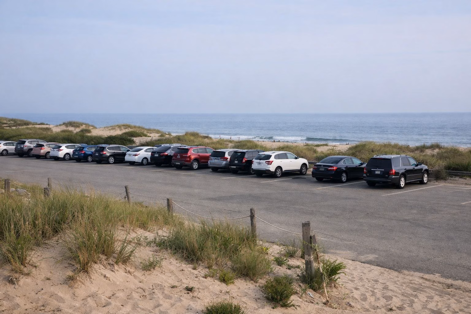 Cars parked in a paved beach lot behind low dunes and beach grass, the ocean beyond