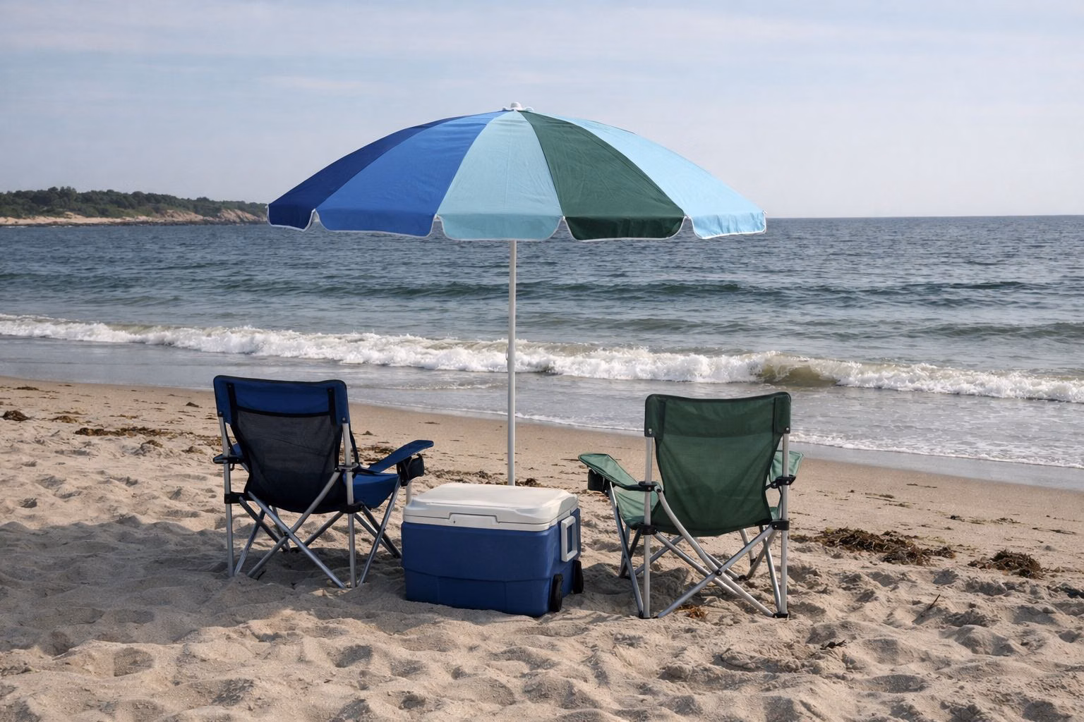 A beach umbrella, two folding chairs and a cooler on the sand near the waterline